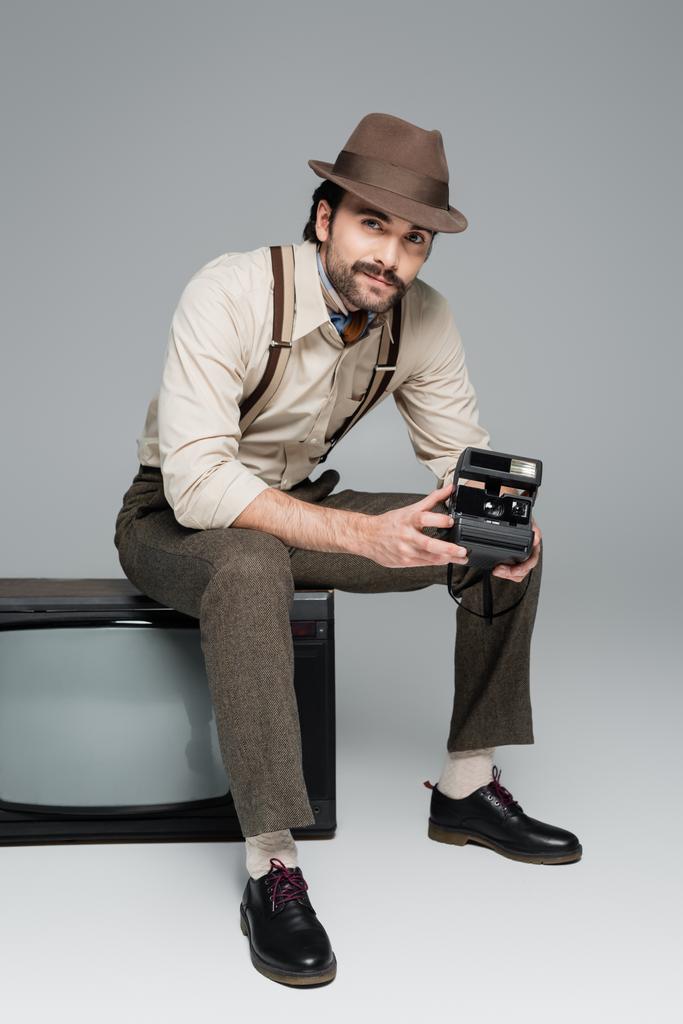 longitud completa de hombre sonriente ropa de estilo retro y sombrero con cámara vintage y sentado en la televisión antigua en gris  - Foto, Imagen