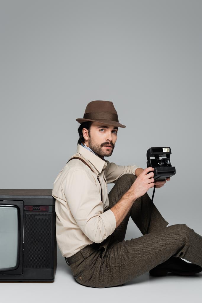 мужчина ретро-стиль одежды и шляпы сидя возле старинного телевизора с винтажной камерой на сером  - Фото, изображение