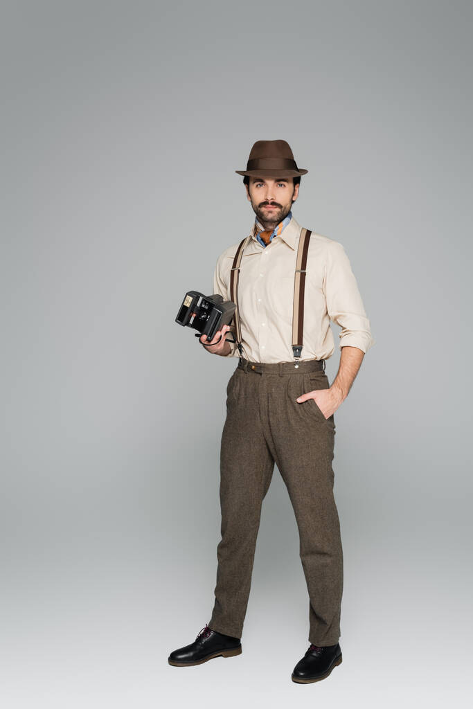 повна довжина чоловіка ретро стиль одяг і капелюх тримає старовинну камеру і стоїть з рукою в кишені на сірому
  - Фото, зображення