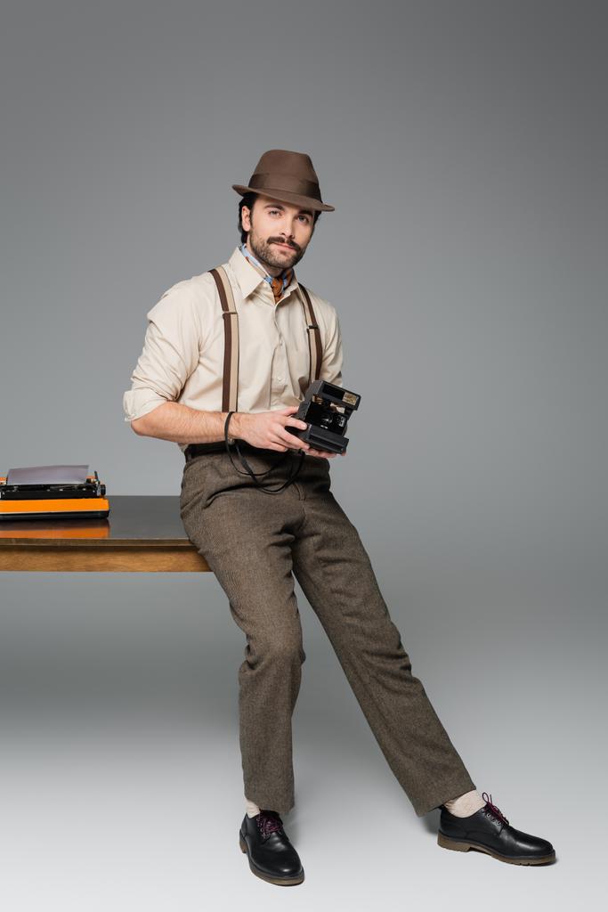 pleine longueur de vêtements de style rétro homme et chapeau tenant caméra vintage et debout près du bureau avec machine à écrire sur gris  - Photo, image