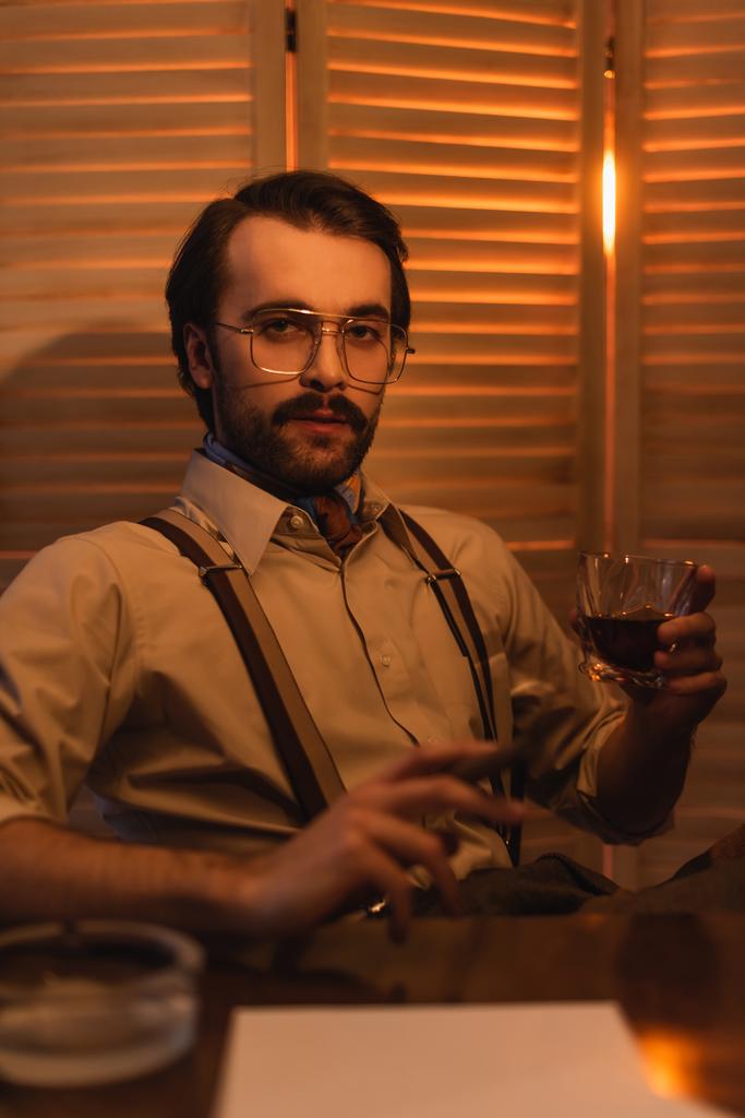 Mann mit Brille hält Glas Whiskey und Zigarre neben verschwommenem Klappbildschirm - Foto, Bild