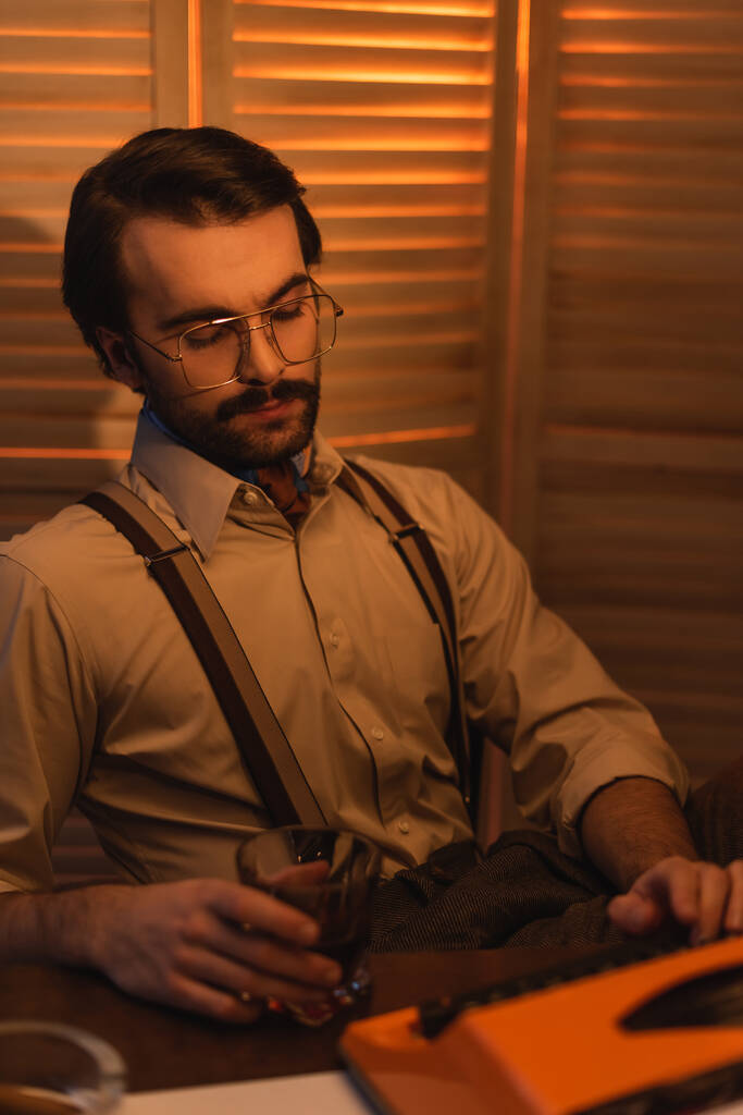 écrivain dans des lunettes tenant un verre de whisky et utilisant une machine à écrire près de l'écran pliant flou - Photo, image