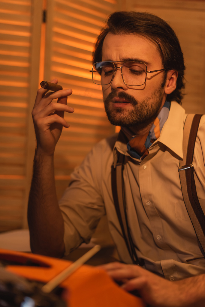δημοσιογράφος με μουστάκι και γυαλιά καπνίζοντας πούρο κοντά σε θολή ρετρό γραφομηχανή  - Φωτογραφία, εικόνα