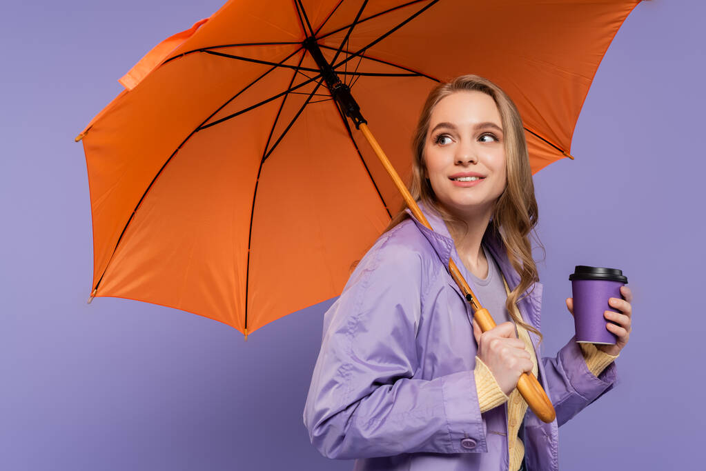 fröhliche junge Frau im Trenchcoat hält Pappbecher in der Hand und steht unter orangefarbenem Regenschirm isoliert auf lila  - Foto, Bild