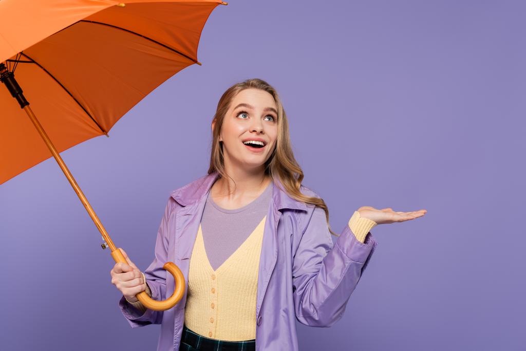 トレンチコート姿の陽気な若い女性が紫に隔離されたオレンジの傘を持ちながらジェスチャー  - 写真・画像