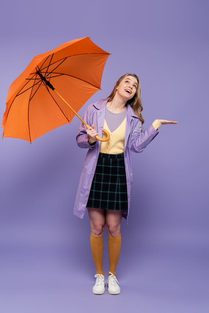 volledige lengte van vrolijke jonge vrouw in trench jas gebaren terwijl het houden van oranje paraplu op paars  - Foto, afbeelding