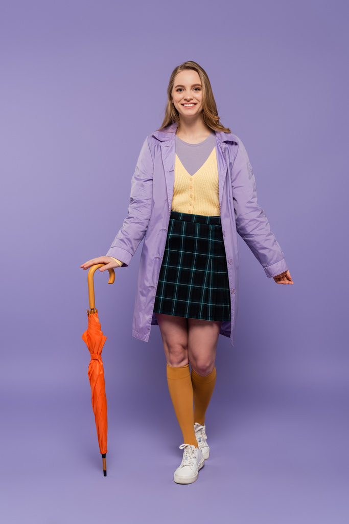 longitud completa de la joven alegre en gabardina de pie con paraguas naranja en púrpura  - Foto, imagen