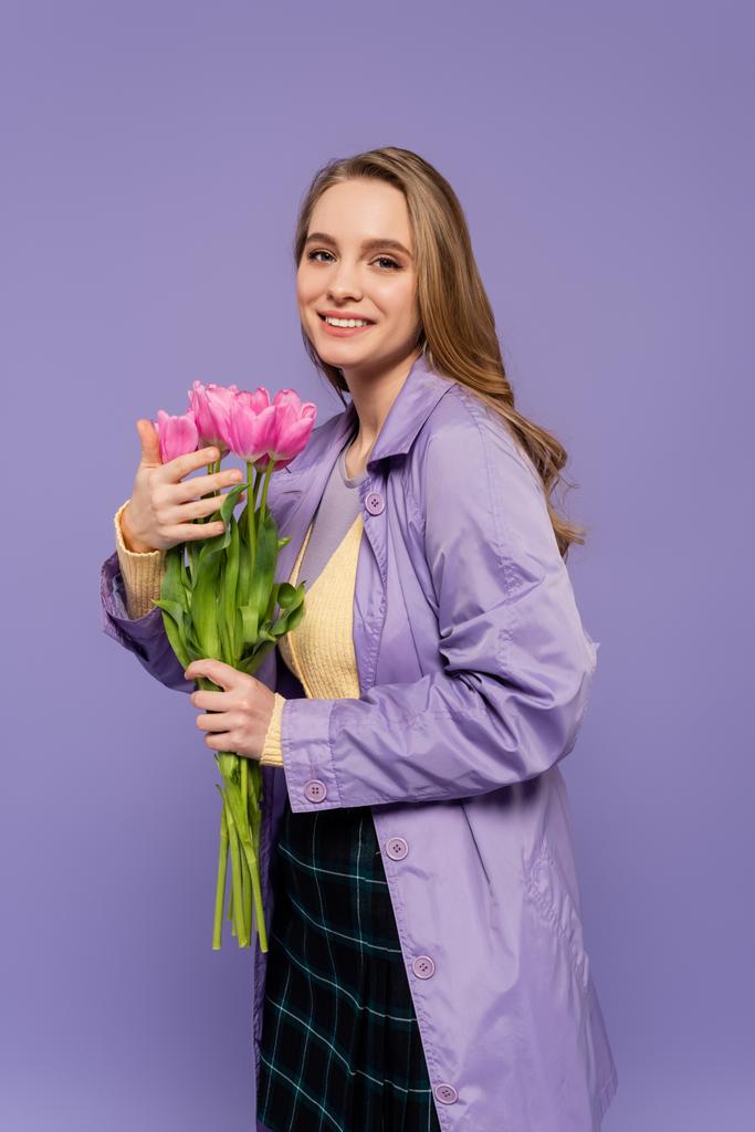 felice giovane donna in trench con tulipani rosa isolati su viola - Foto, immagini