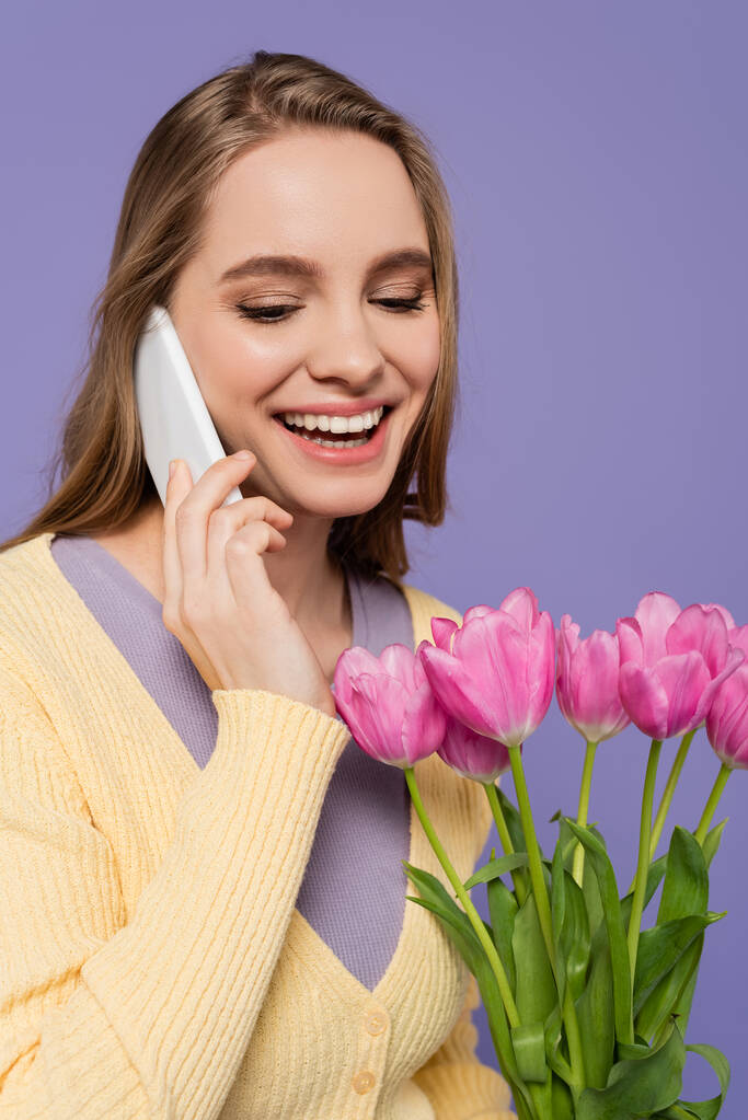 jovem alegre segurando tulipas rosa e falando no smartphone isolado em roxo - Foto, Imagem