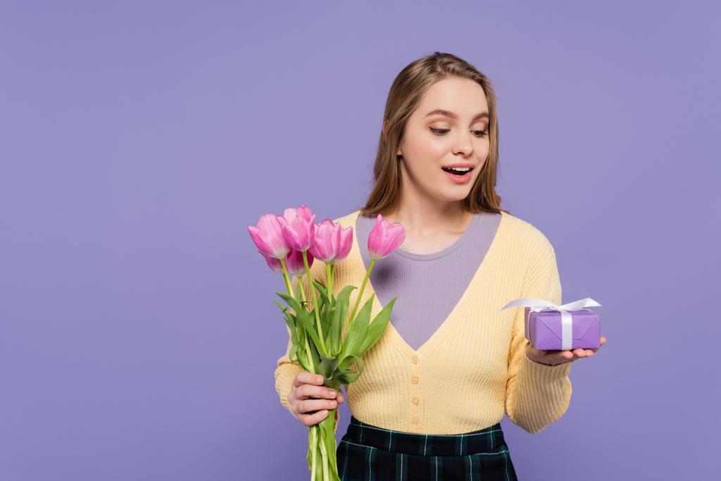 jovem mulher surpreso segurando tulipas rosa e caixa de presente isolado em roxo - Foto, Imagem