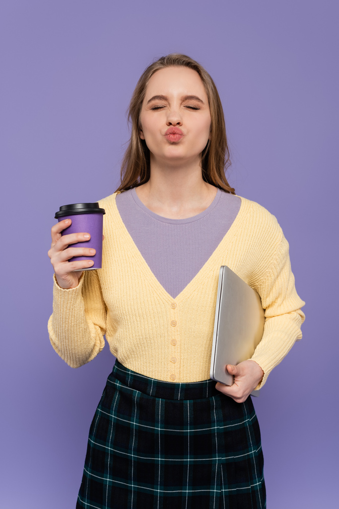 молодая женщина дуть губы, держа ноутбук и бумажный стаканчик изолирован на фиолетовый  - Фото, изображение