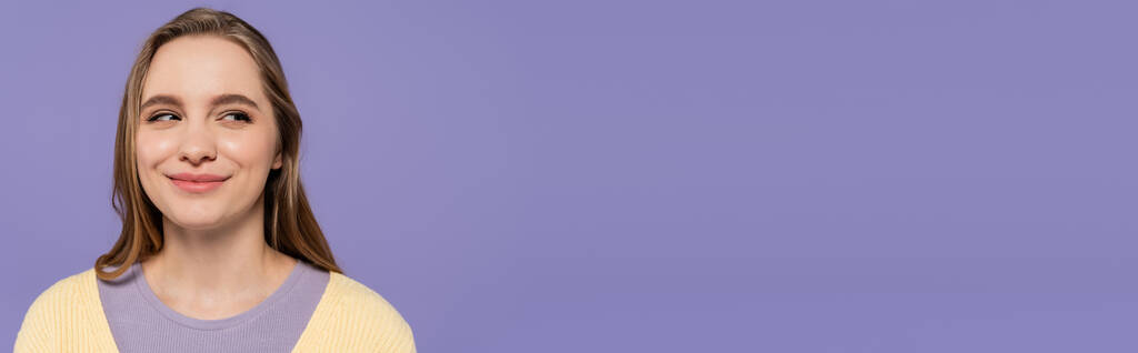 sluwe jonge vrouw kijken weg en glimlachen geïsoleerd op paars, banner - Foto, afbeelding