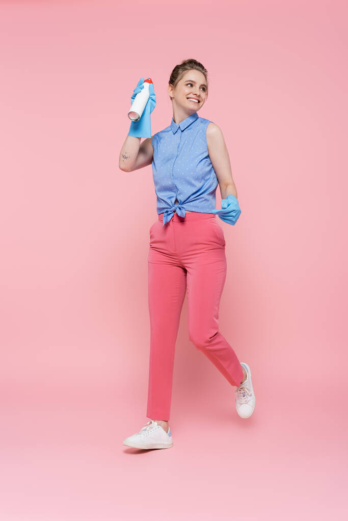 volledige lengte van gelukkig jonge vrouw in rubber handschoenen houden luchtverfrisser fles op roze  - Foto, afbeelding