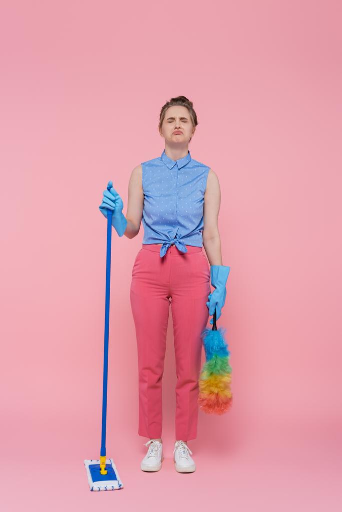 plná délka rozrušená mladá žena v gumových rukavicích drží mop a prach kartáč na růžové - Fotografie, Obrázek