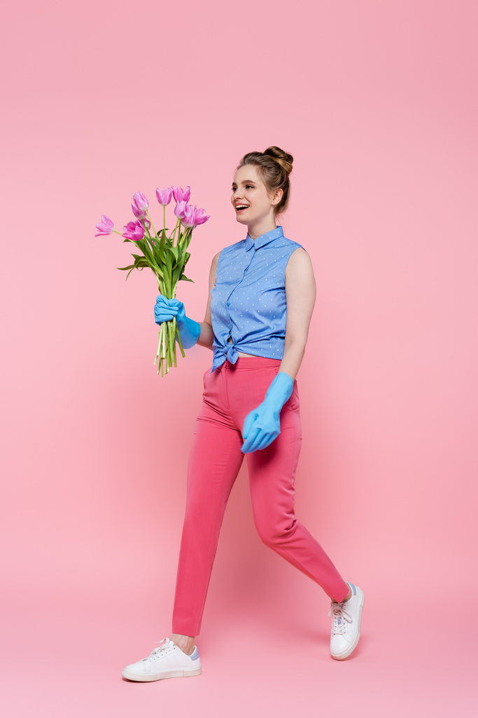 volledige lengte van gelukkig jong vrouw in rubber handschoenen houden tulpen en lopen op roze - Foto, afbeelding
