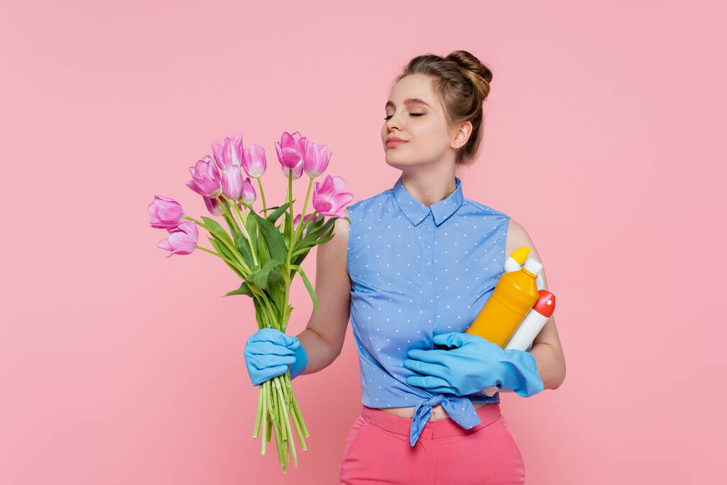 tevreden jonge vrouw in rubber handschoenen houden flessen met wasmiddel en ruikende tulpen geïsoleerd op roze - Foto, afbeelding