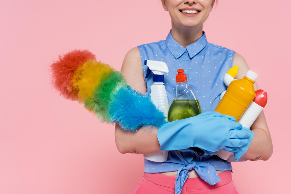 przycięty widok zadowolonej młodej kobiety w gumowych rękawiczkach trzymających butelki z detergentem i szczotką do kurzu izolowane na różowy - Zdjęcie, obraz