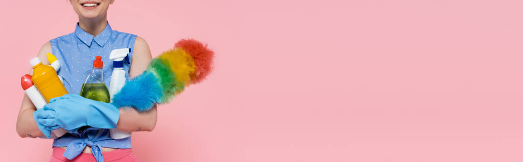 vista recortada de la joven complacida en guantes de goma sosteniendo botellas con detergente y cepillo de polvo aislado en rosa, pancarta - Foto, Imagen