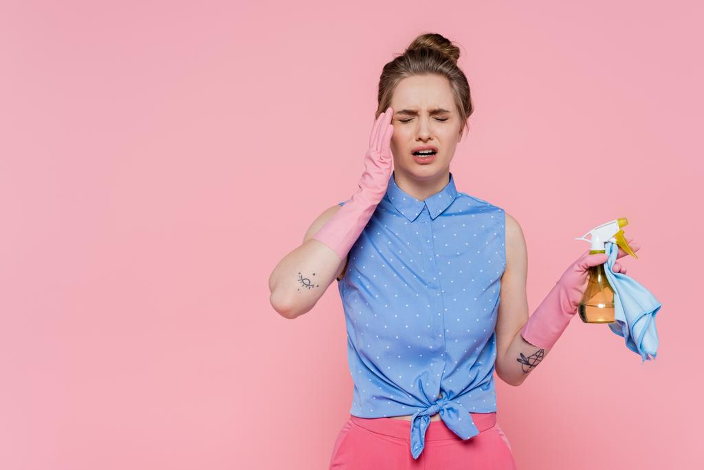 wyczerpana młoda kobieta w gumowych rękawiczkach trzymająca niebieską szmatę i butelkę z rozpylaczem na różowo  - Zdjęcie, obraz
