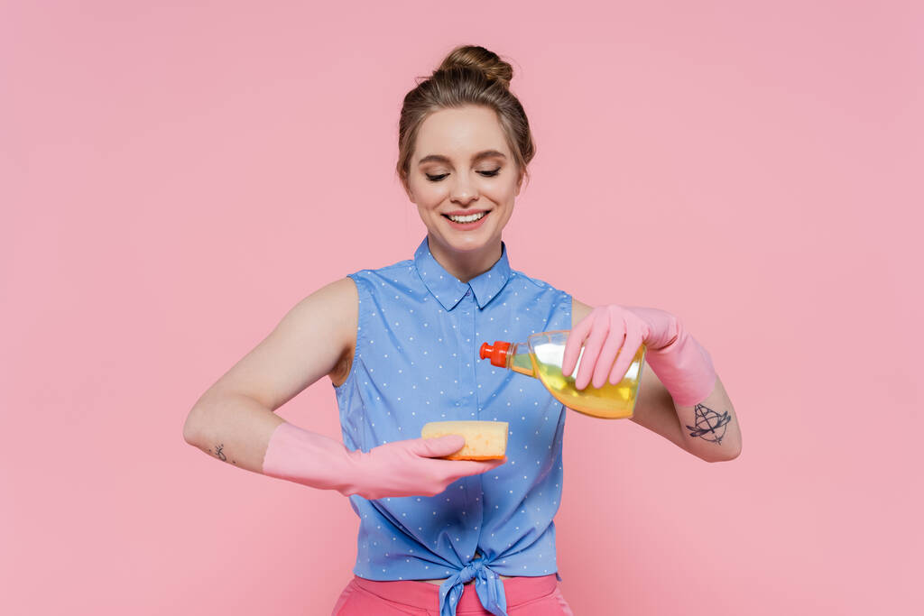 τατουάζ νεαρή γυναίκα κρατώντας σφουγγάρι και μπουκάλι με απορρυπαντικό, ενώ χαμογελά απομονωμένη σε ροζ  - Φωτογραφία, εικόνα