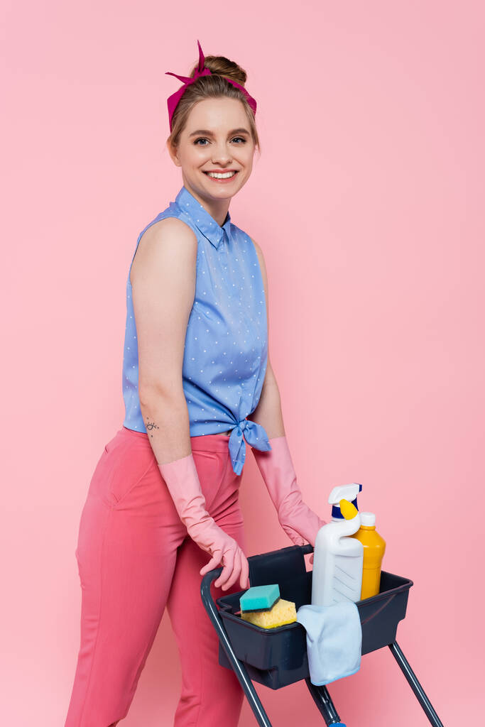heureuse jeune femme en gants de caoutchouc debout près du chariot d'entretien ménager avec des fournitures de nettoyage isolé sur rose - Photo, image
