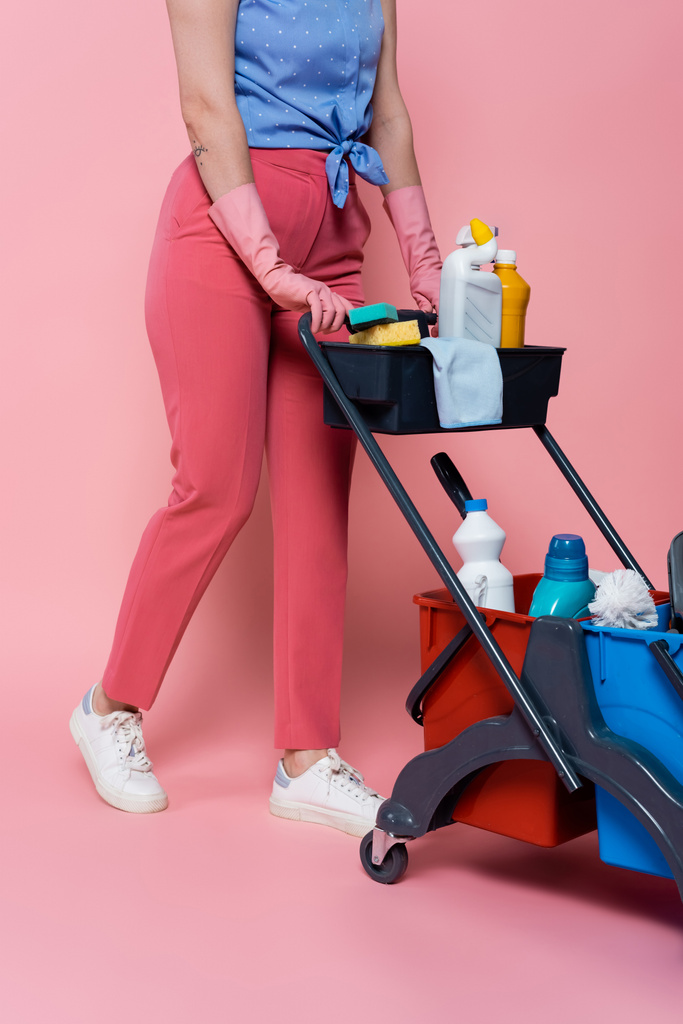 przycięty widok wytatuowanej kobiety w gumowych rękawiczkach stojącej w pobliżu wózka sprzątającego z dodatkami do czyszczenia na różowo - Zdjęcie, obraz