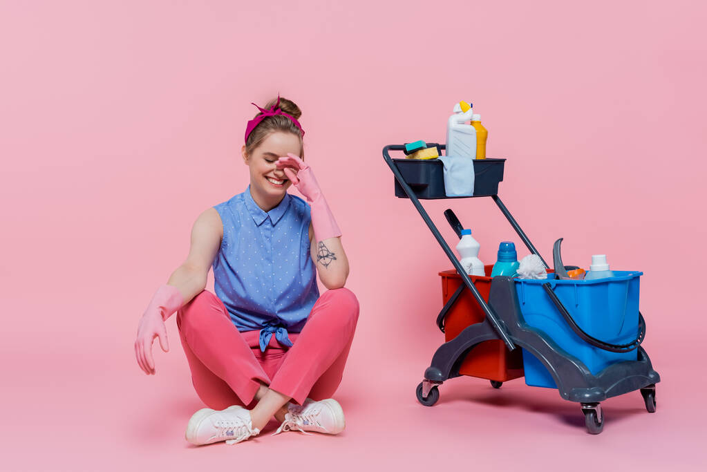 longitud completa de la joven sonriente en guantes de goma sentado cerca del carro de limpieza con suministros de limpieza en rosa - Foto, imagen