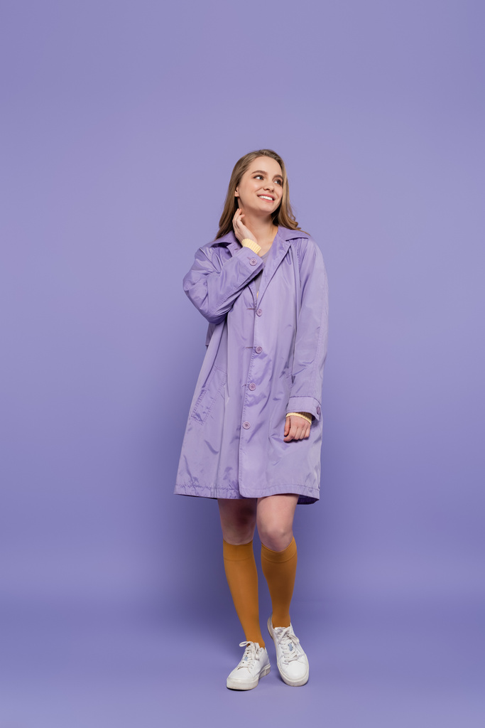 volledige lengte van gelukkig jonge vrouw in violette regenjas staan op paars - Foto, afbeelding