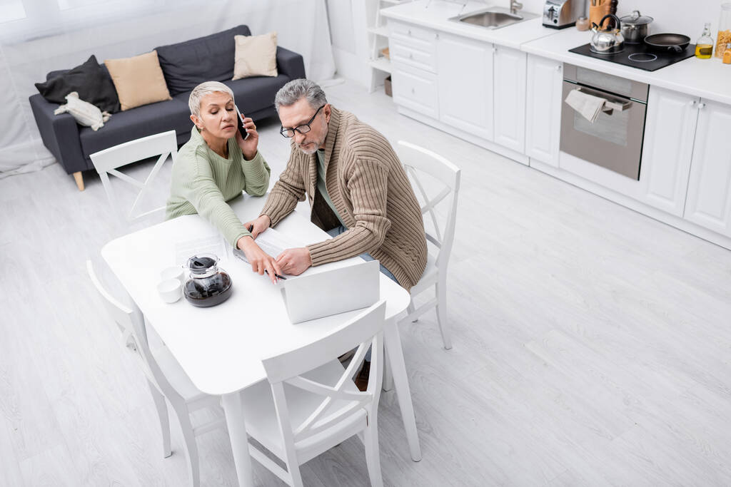Vista aérea de la mujer mayor hablando por teléfono celular y apuntando a la computadora portátil cerca del marido con papel en la cocina  - Foto, Imagen