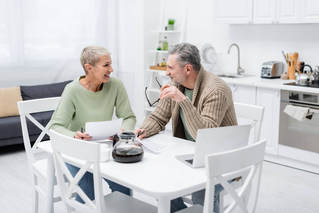 Allegro coppia anziana parlando vicino a carte e laptop in cucina  - Foto, immagini