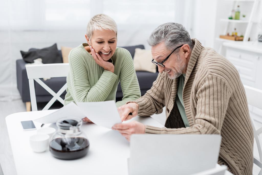 Positive pensioner pointing at paper near wife and devices in kitchen  - Photo, Image