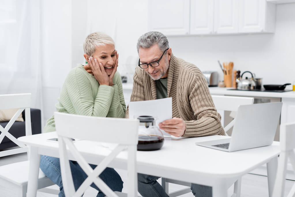 Cheerful senior woman looking at document near husband and laptop in kitchen  - Photo, Image