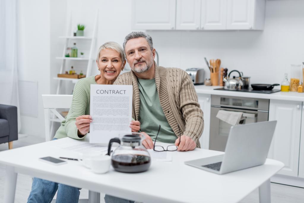 Усміхнена старша жінка тримає контракт біля чоловіка і пристроїв на кухні
  - Фото, зображення