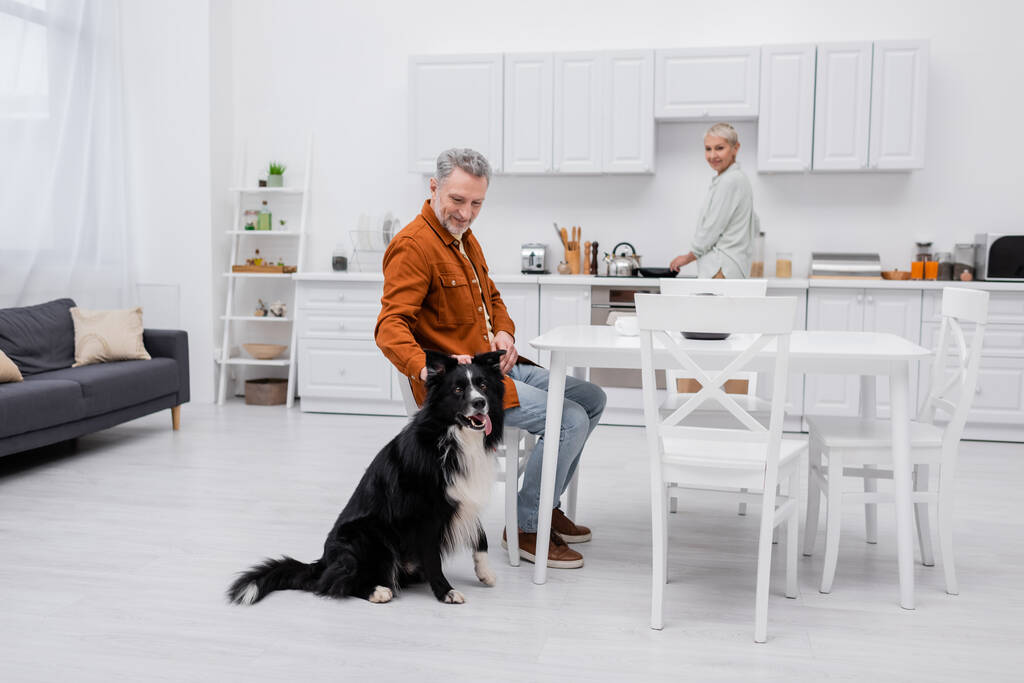 Neşeli, olgun bir adam, mutfakta karısının yanında çoban köpeği besliyor.  - Fotoğraf, Görsel