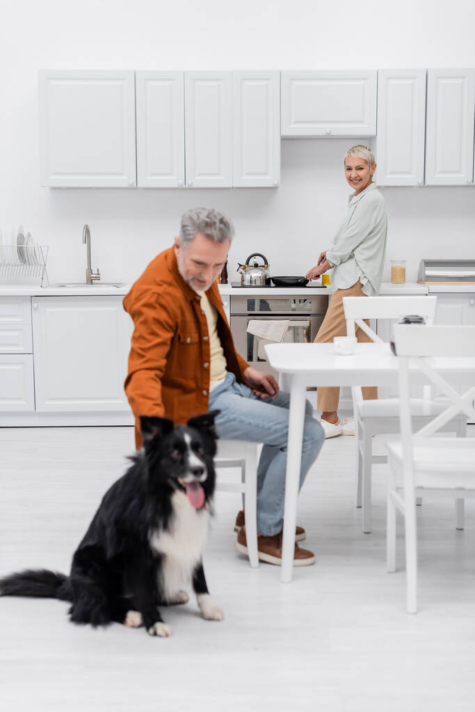 Щаслива старша жінка готує біля розмитого чоловіка погладжуючи кордон колі на кухні
  - Фото, зображення
