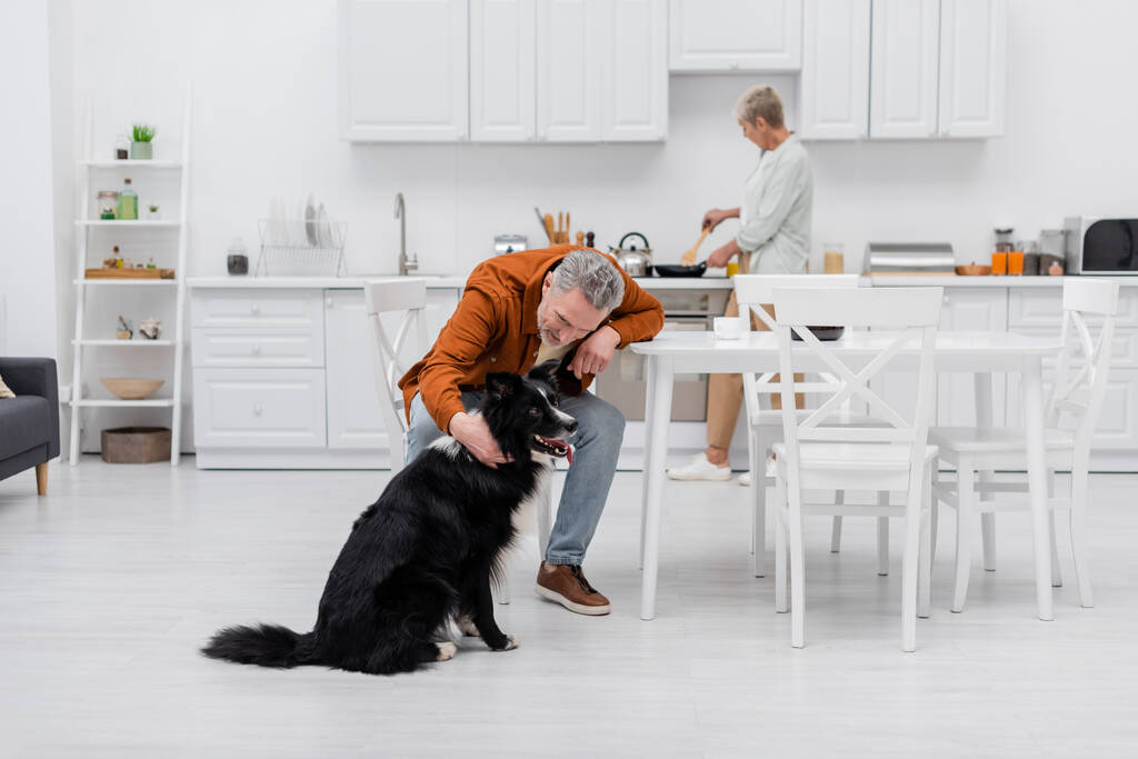 Mann mittleren Alters streichelt Border Collie neben Kaffeetasse in Küche  - Foto, Bild
