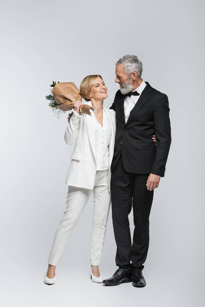 Positivo maturo sposa tenendo bouquet di nozze e abbracciare lo sposo su sfondo grigio  - Foto, immagini