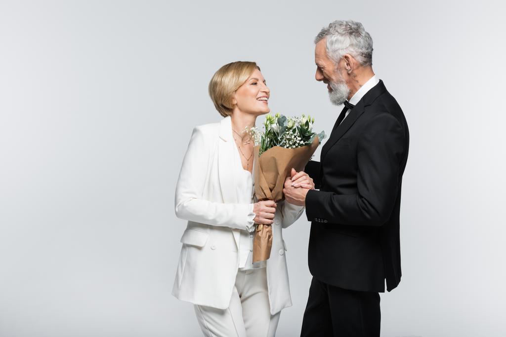 Positiv reife Braut hält Strauß und Hand des Bräutigams isoliert auf grau  - Foto, Bild