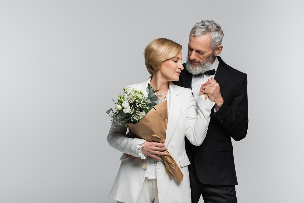 Μεσήλικας γαμπρός κρατώντας το χέρι της νύφης με γαμήλιο μπουκέτο απομονώνονται σε γκρι  - Φωτογραφία, εικόνα
