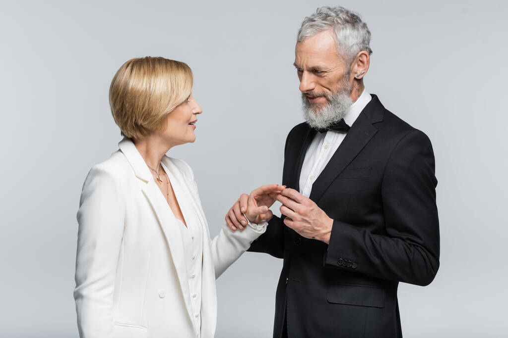 Middle aged groom wearing wedding ring on smiling bride isolated on grey  - Photo, Image