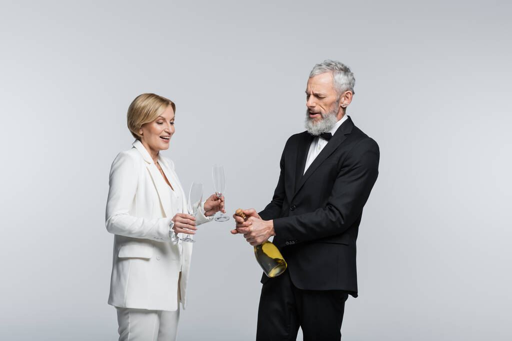 Starší ženich otevření šampaňské poblíž pozitivní nevěsty s brýlemi izolované na šedé  - Fotografie, Obrázek