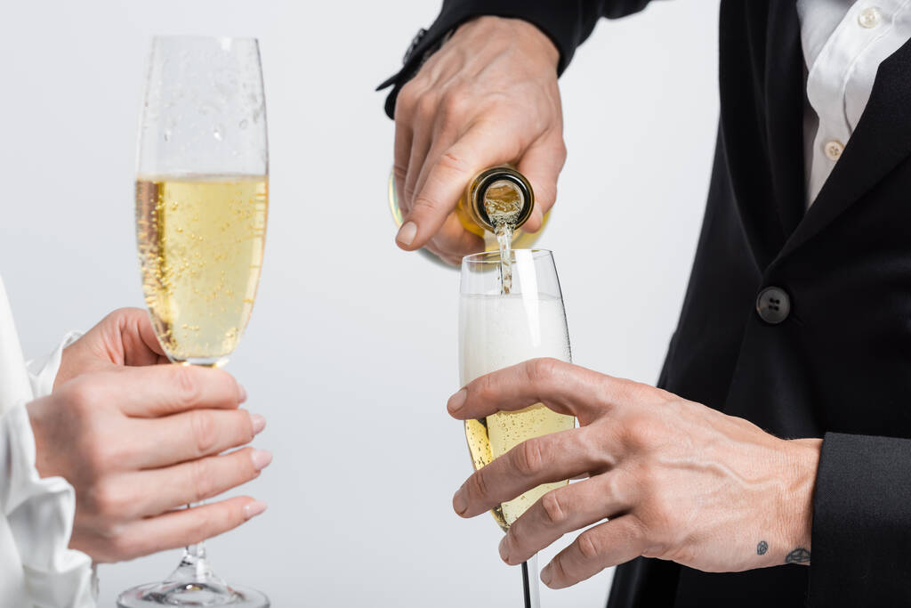 Обрезанный вид жениха, наливающего шампанское рядом с невестой со стаканом, изолированным на сером  - Фото, изображение