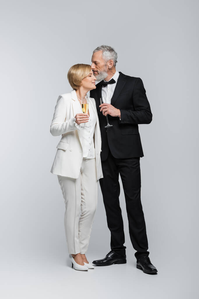 Marié d'âge moyen tenant un verre de champagne et embrassant mariée sur fond gris  - Photo, image