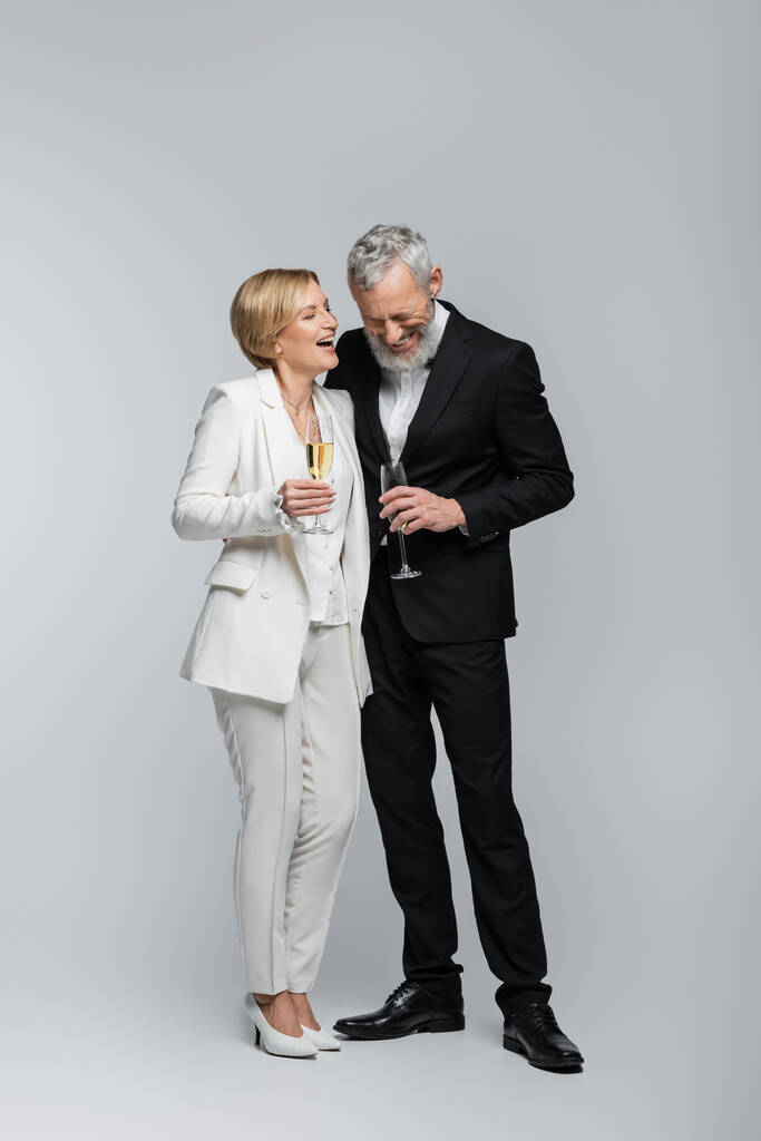 Glückliche reife Braut mit einem Glas Champagner in der Nähe des Bräutigams im Anzug auf grauem Hintergrund  - Foto, Bild