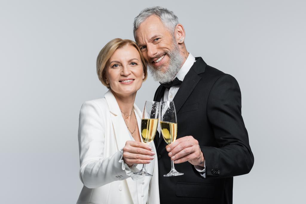 Усміхнений зрілий наречений дивиться на камеру біля нареченої з шампанським ізольовано на сірому
  - Фото, зображення