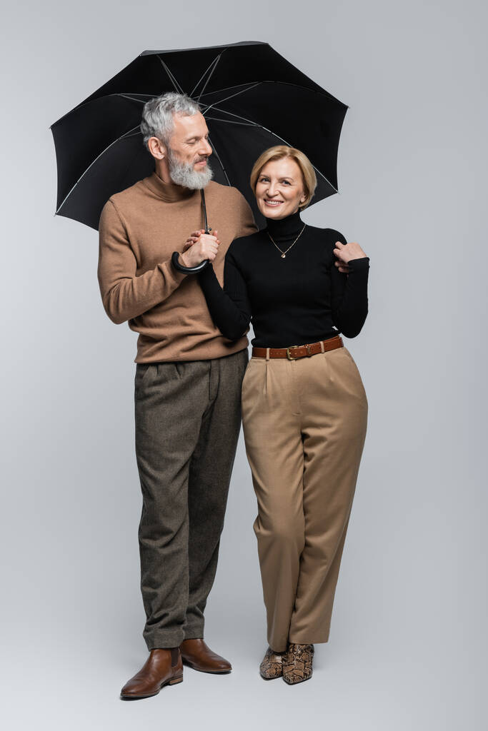 Θετική ώριμη γυναίκα κοιτάζοντας κάμερα κοντά κομψό σύζυγο με ομπρέλα σε γκρι φόντο  - Φωτογραφία, εικόνα