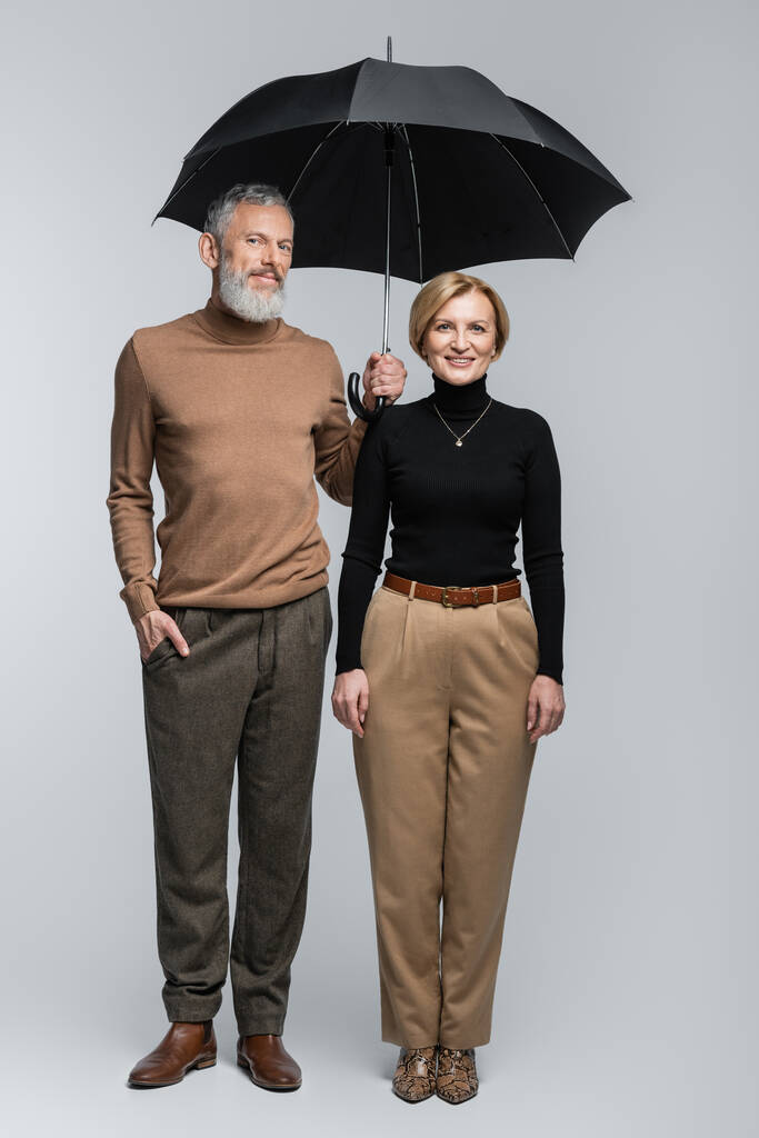 Pleine longueur de couple mature élégant avec parapluie regardant la caméra sur fond gris  - Photo, image