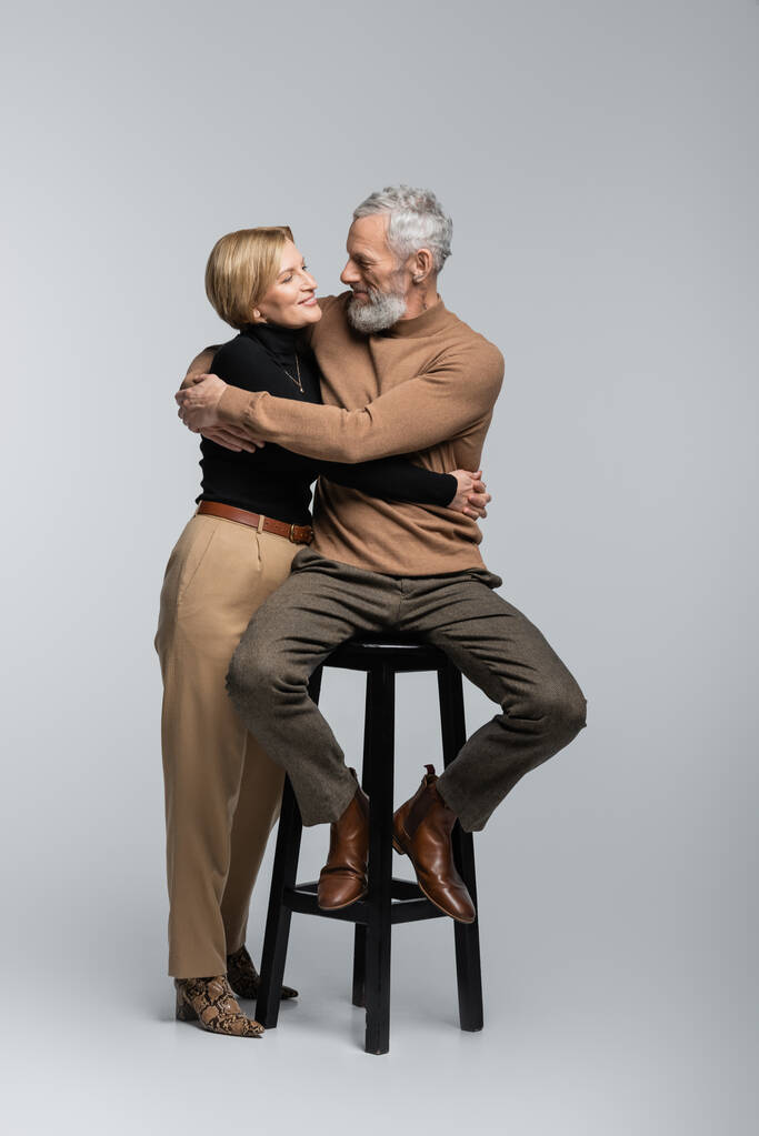 Reife Frau umarmt stilvollen Ehemann sitzt auf Stuhl auf grauem Hintergrund  - Foto, Bild
