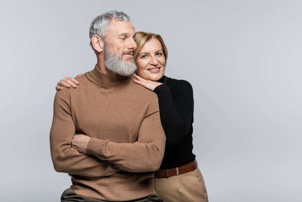 Usmívající se starší žena v roláku objímající manžela a při pohledu na kameru izolované na šedé  - Fotografie, Obrázek
