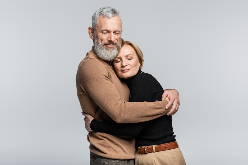 Bearded grey haired man hugging wife with closed eyes isolated on grey  - Photo, Image